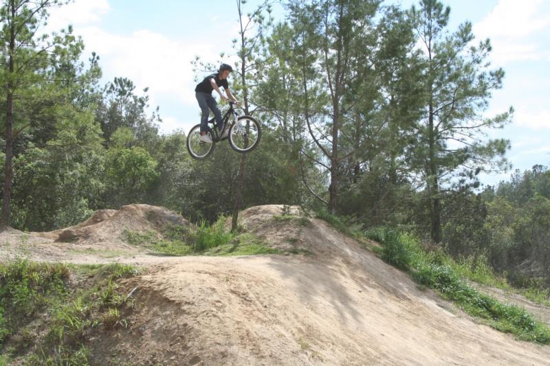 A person riding a bicycle is airborne above a dirt ramp in a natural environment, surrounded by trees and greenery. The rider is wearing a helmet and casual clothing, showcasing a BMX biking trick. Santos mountain bike trail.