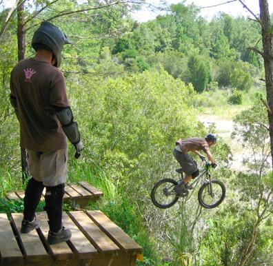 A mountain biker performs a jump off a wooden platform, with a person in protective gear observing from the side. The background features lush greenery and trees, indicating an outdoor setting. Santos mountain bike trail.