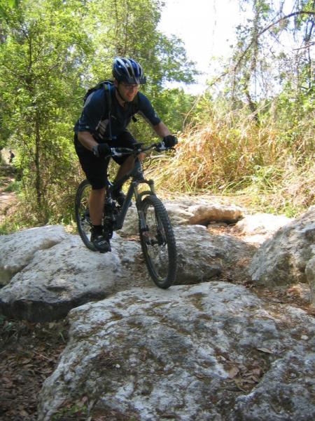 A mountain biker navigating rocky terrain on a forest trail, wearing a helmet and cycling gear. The background features lush greenery and scattered boulders, showcasing an outdoor adventure setting. Santos mountain bike trail.