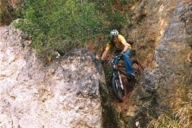A mountain biker navigating a narrow rocky descent surrounded by tall grass and trees, wearing a helmet and a yellow shirt. In the background, other people are watching from a higher vantage point. Santos mountain bike trail.