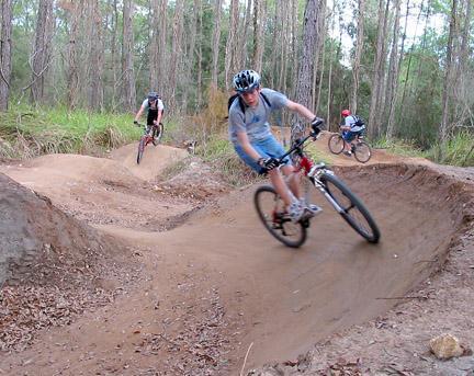 Mountain bikers navigating a dirt trail with curves in a forested area, surrounded by trees and underbrush. Santos mountain bike trail.