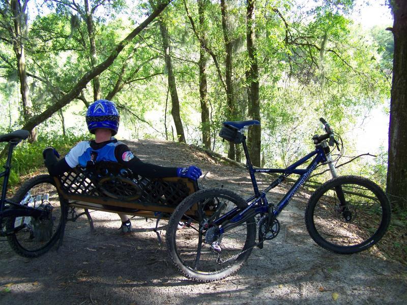 A young cyclist wearing a blue helmet and jersey relaxes on a wooden bench beside a mountain bike. The scene is set along a wooded path, surrounded by green trees and foliage, with a glimpse of water in the background. Balm Boyette Scrub Preserve mountain bike trail.