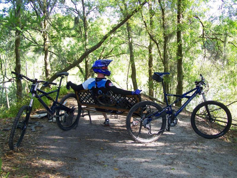 A person resting on a bench in a forested area, wearing a blue helmet and cycling gear, with two mountain bikes parked nearby. Sunlight filters through the trees, creating a serene outdoor setting. Balm Boyette Scrub Preserve mountain bike trail.