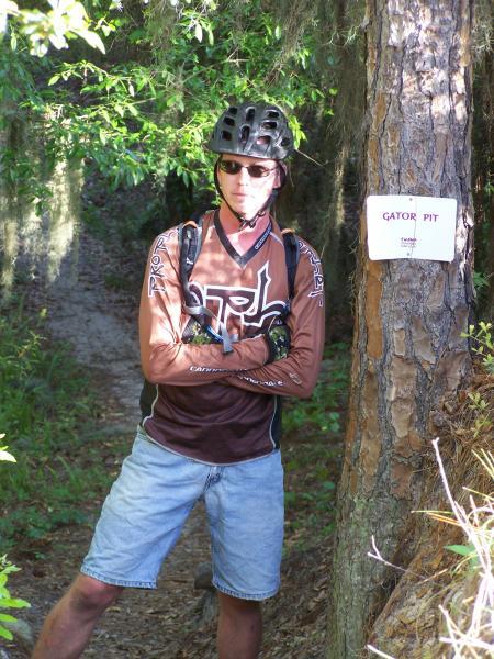 A mountain biker wearing a helmet and sunglasses stands confidently with arms crossed beside a tree. A sign labeled "GATOR PIT" is attached to the tree, indicating the entrance to a biking trail. The surrounding area features lush greenery, suggesting a forested setting. Balm Boyette Scrub Preserve mountain bike trail.