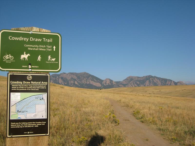 Signage marking the Cowdrey Draw Trail with directions for biking, horseback riding, and hiking. The background features open fields and mountain ranges under a clear blue sky. Marshall Road Open Spaces mountain bike trail.