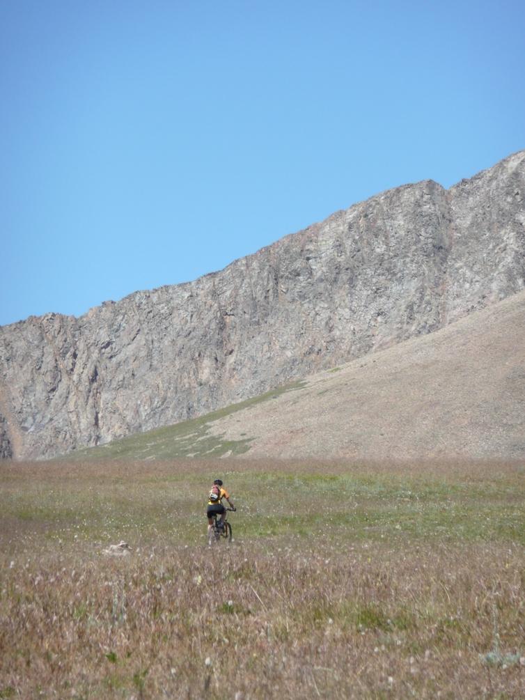 A cyclist riding a mountain bike across a grassy field with a rocky hillside and clear blue sky in the background. Colorado Trail: Kenosha Pass To Breckenridge mountain bike trail.