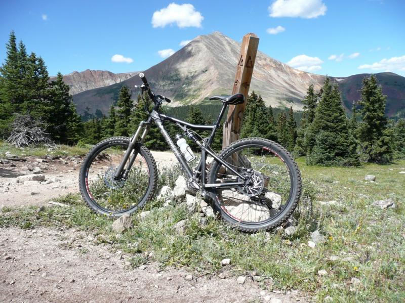 A mountain bike resting against a rock pile on a hiking trail, with a backdrop of a mountain and lush green trees. The sky is bright blue with scattered clouds. A trail sign is visible nearby, indicating the outdoor setting. Colorado Trail: Kenosha Pass To Breckenridge mountain bike trail.