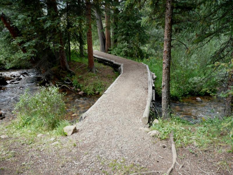 A narrow, winding path made of gravel stretches through a lush forest, bordered by tall trees. On one side, a clear stream flows peacefully, while greenery and small plants line the edges of the trail. The scene captures a serene and natural outdoor environment. Colorado Trail: Kenosha Pass To Breckenridge mountain bike trail.