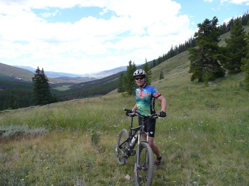 A mountain biker stands beside his bike on a grassy hillside, surrounded by a scenic landscape of rolling hills and pine trees under a partly cloudy sky. The biker is wearing a colorful cycling jersey and a helmet, smiling as he enjoys the outdoors. Colorado Trail: Kenosha Pass To Breckenridge mountain bike trail.