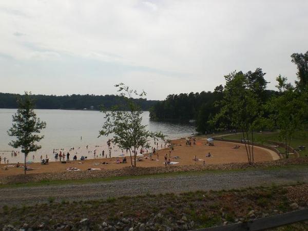 A scenic view of a sandy beach by a lake, with several people enjoying the water and sunbathing. The shoreline is lined with trees, and a grassy area can be seen nearby. The sky is overcast, suggesting a calm day for leisure activities. Itusi @ Lake Norman State Park mountain bike trail.