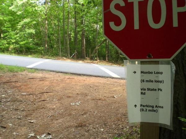 A stop sign next to a dirt path leading to a road, with a sign below it indicating directions for the Monbo Loop trail (6-mile loop) and a parking area (0.2 mile). The scene is set in a lush, green forest. Itusi @ Lake Norman State Park mountain bike trail.