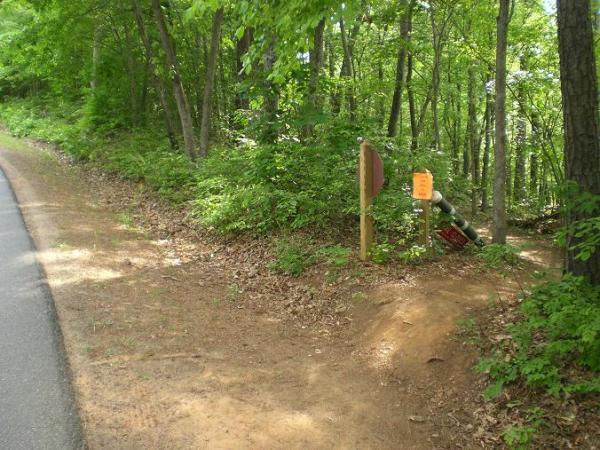 A dirt path leading into a wooded area next to a paved road. A wooden signpost is visible, with a brown sign and an orange notice attached, indicating a trail or area of interest. Surrounding vegetation is lush and green, showcasing a bright, sunny day. Itusi @ Lake Norman State Park mountain bike trail.