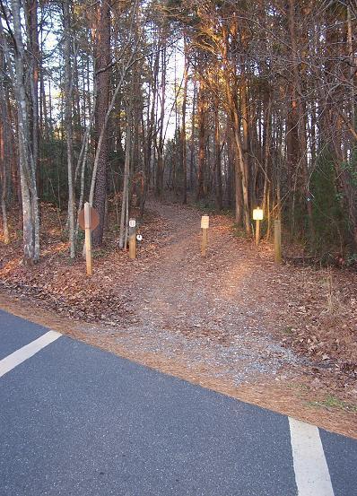 A gravel path leading into a wooded area, lined with trees. At the entrance of the path, there are several wooden posts with signs, including a round trail marker and informational signs. The surrounding ground is covered with fallen leaves, and part of a paved road is visible at the bottom of the image. The scene is illuminated by soft, natural light, suggesting early morning or late afternoon. Itusi @ Lake Norman State Park mountain bike trail.