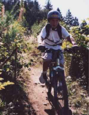 A young boy riding a mountain bike on a narrow trail surrounded by greenery and trees on a sunny day. He is wearing a helmet and a backpack, focused on navigating the path ahead. Galbraith Mountain mountain bike trail.