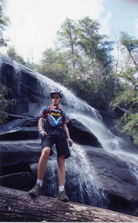 A person in cycling attire stands on a log near a rocky waterfall, surrounded by lush green foliage. The individual is wearing a helmet and sunglasses, with water cascading down the rocks behind them under a partly cloudy sky. Daniel Ridge mountain bike trail.