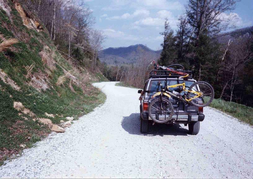 A gravel road curving through a wooded area with mountains in the background. A vehicle is parked on the side of the road, featuring two bicycles secured on a rack on its roof and one attached to the back. The scene is set under a clear blue sky, with patches of green grass and scattered foliage along the roadside. Daniel Ridge mountain bike trail.