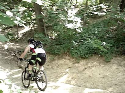 A person riding a mountain bike downhill on a dirt trail surrounded by lush greenery and trees. The cyclist is wearing a helmet and a colorful outfit, and appears focused on navigating the path. Rowlett Creek Preserve mountain bike trail.