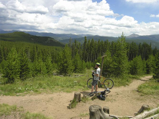A mountain biker stands beside their bike on a wooded trail, gazing out at a panoramic view of rolling hills and dense forests under a partly cloudy sky. Peaks Trail mountain bike trail.