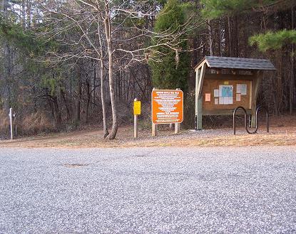 Image of a trailhead area with a wooden information kiosk housing a map and local guidelines. There's an orange sign with instructions or important information nearby, as well as a bike rack. The background features trees and a gravel path leading into a wooded area. Itusi @ Lake Norman State Park mountain bike trail.
