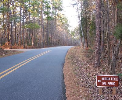 A winding road surrounded by trees with a sign indicating the direction to mountain bicycle trail parking. The scene captures a serene outdoor setting. Itusi @ Lake Norman State Park mountain bike trail.