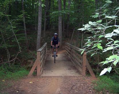 A cyclist riding a mountain bike across a wooden bridge surrounded by dense green trees in a forest setting. Itusi @ Lake Norman State Park mountain bike trail.