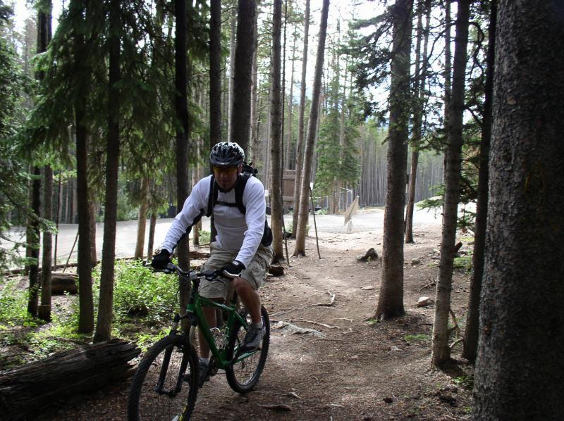A mountain biker riding a green bicycle along a narrow trail in a forested area, surrounded by tall trees and natural greenery. The cyclist is wearing a white long-sleeve shirt, shorts, and a helmet, with a backpack attached. In the background, there is a pathway leading to a clearing. Peaks Trail mountain bike trail.