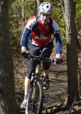 A mountain biker navigating a dirt trail surrounded by trees, wearing a red and blue cycling jersey, a helmet, and shorts. The biker is focused as they ride over uneven terrain. Gambrill State Park mountain bike trail.
