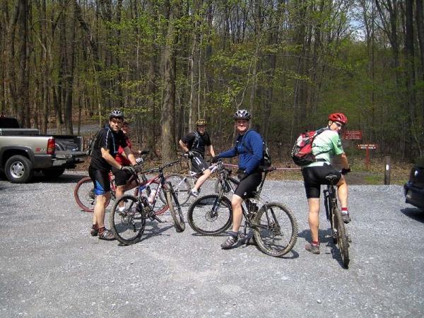 Five mountain bikers are gathered in a gravel parking area surrounded by trees. They are wearing cycling gear and helmets, and each is standing beside their bike. Some are looking at the camera, while others are engaged in conversation. In the background, parked vehicles and trail signs can be seen, indicating a recreational area. Gambrill State Park mountain bike trail.