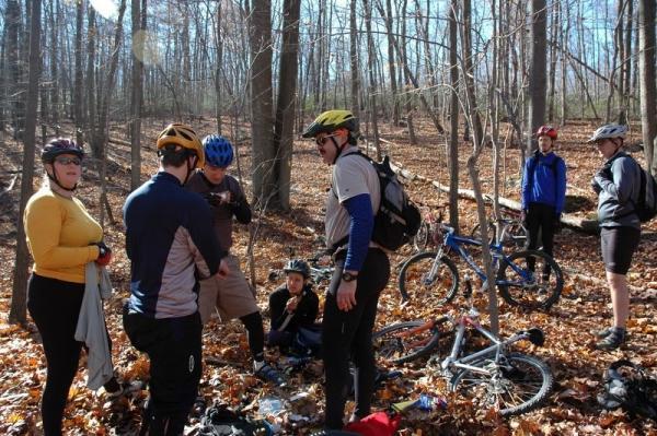 A group of eight cyclists gathered in a forest during autumn. They are wearing helmets and cycling gear, and some are crouched down, engaged in a discussion or inspecting equipment. Several bicycles are parked nearby on the ground, surrounded by fallen leaves and trees without foliage in the background. The scene captures a moment of collaboration or a break during their biking adventure. Gambrill State Park mountain bike trail.