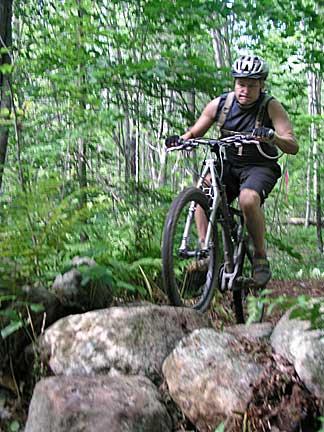 A mountain biker navigating over a rocky trail in a dense green forest. The cyclist is wearing a helmet and performance gear, showcasing outdoor biking in a natural setting. Bradbury Mt State Park mountain bike trail.