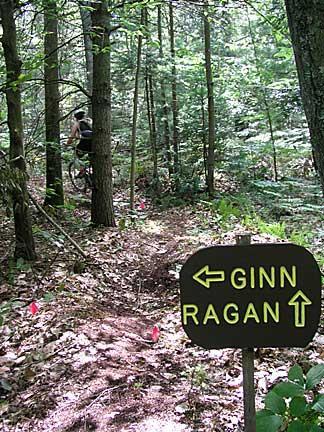 A trail sign in a wooded area, indicating directions with arrows pointing left toward "GINN" and straight ahead toward "RAGAN." The path is lined with small flags and surrounded by dense greenery and trees, suggesting a hiking or biking trail. Bradbury Mt State Park mountain bike trail.