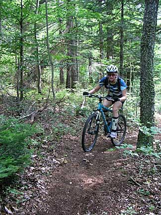 A person riding a mountain bike along a forest trail surrounded by greenery, with tall trees and plants in the background. The cyclist is wearing a helmet and cycling gear. Bradbury Mt State Park mountain bike trail.