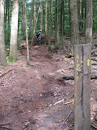 A mountain bike trail in a forested area, featuring a wooden sign indicating the path ahead. A cyclist is visible in the background, riding along the dirt trail surrounded by trees and foliage. Bradbury Mt State Park mountain bike trail.