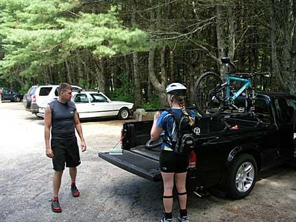A man and a woman prepare for a biking trip in a forested parking area. The man stands beside a black pickup truck, while the woman, wearing a bike helmet and athletic gear, is seen checking the contents of the truck bed that holds a bicycle. Several parked vehicles and trees are visible in the background. Bradbury Mt State Park mountain bike trail.