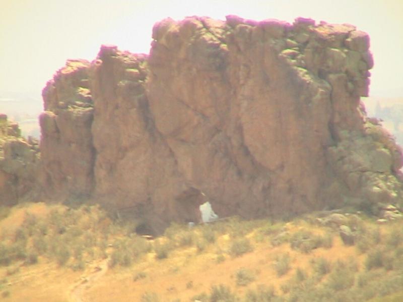 A large, rugged rock formation with a natural arch, surrounded by sparse vegetation and open terrain under a hazy sky. Devil's Backbone mountain bike trail.