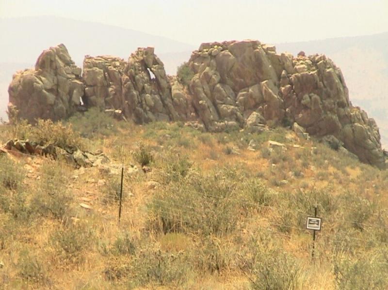 Rock formations on a hillside surrounded by dry vegetation and shrubs, with a sign visible in the foreground. The background features a hazy landscape of rolling hills under a pale sky. Devil's Backbone mountain bike trail.