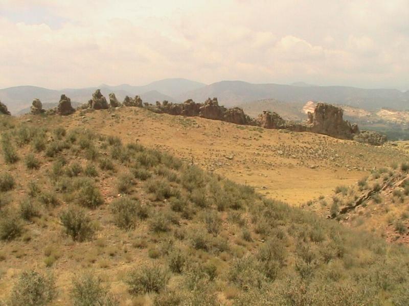 A panoramic view of a rocky landscape with rolling hills covered in sparse vegetation, under a partly cloudy sky. The foreground features patches of low shrubs, while a rugged outcrop of rocks is visible in the midground, leading to distant mountains on the horizon. Devil's Backbone mountain bike trail.