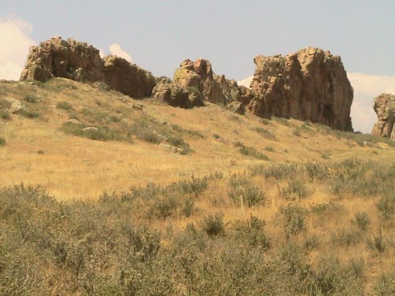 Rock formations on a grassy hillside under a blue sky with scattered clouds. The landscape features dry grass and sparse vegetation in the foreground, with rugged cliffs rising in the background. Devil's Backbone mountain bike trail.