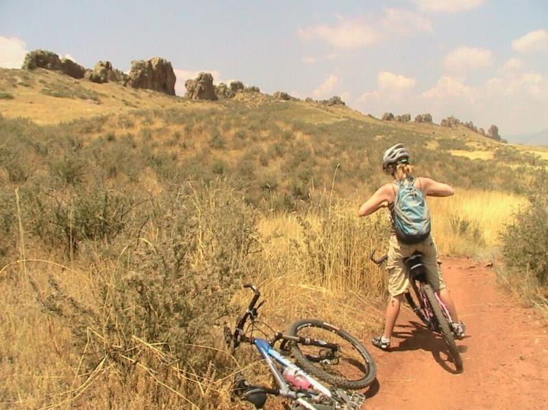 A person in a helmet and backpack stands beside a mountain bike on a dirt trail surrounded by grassy fields and rocky formations in the background. The landscape is sunny with a few clouds in the sky. Devil's Backbone mountain bike trail.