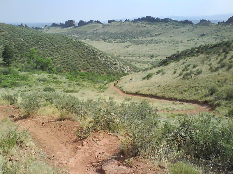 A winding dirt trail surrounded by green hills under a clear sky. The landscape features sparse vegetation and rocky outcrops in the distance, creating a serene outdoor scene suitable for hiking or nature exploration. Devil's Backbone mountain bike trail.