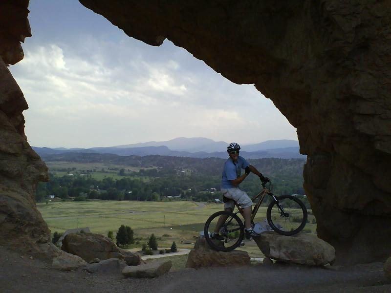 A person standing on a mountain bike poses at the edge of a rock formation, framed by an archway of stone. In the background, a panoramic view of rolling hills and mountains is visible under a cloudy sky. The landscape includes patches of green fields and trees. Devil's Backbone mountain bike trail.