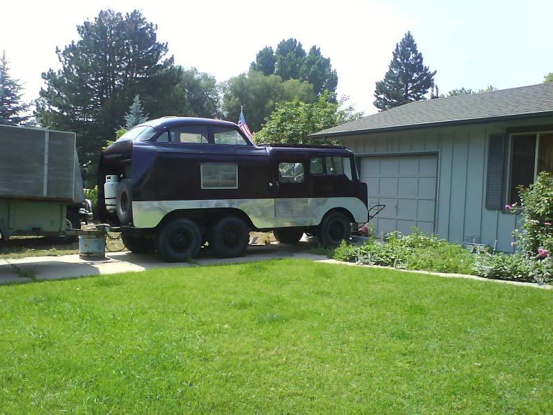 A retro-style camper vehicle with a rounded top, parked on a driveway surrounded by green grass and flower beds. In the background, a single-story house with gray siding and a garage door, along with trees and shrubs in the area. An American flag is visible on the camper. Devil's Backbone mountain bike trail.
