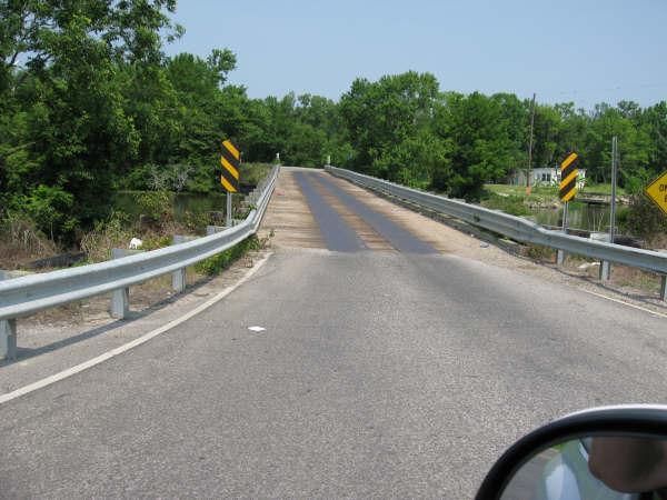 A view of a bridge with a gravel surface, surrounded by greenery, and flanked by safety barriers and warning signs. The sky is clear, indicating a bright day. The photo is taken from a vehicle perspective. Lake Fausse Pointe Trail System mountain bike trail.