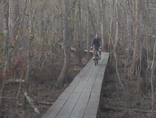A person riding a bicycle on a wooden boardwalk pathway through a dense forest with bare trees. The scene captures a serene, natural setting, highlighting the tranquility of outdoor exploration. Lake Fausse Pointe Trail System mountain bike trail.