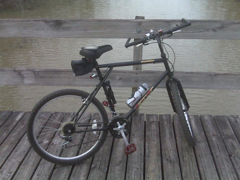 A black mountain bike parked on a wooden deck, with a body of water visible in the background. The bike features thick tires, a water bottle holder, and has a small bag attached to the seat. Lake Fausse Pointe Trail System mountain bike trail.
