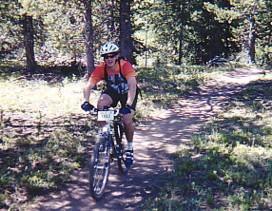 A mountain biker riding along a dirt trail in a forested area, wearing a helmet and bright orange shirt, with a backpack and cycling gear. Sunlight filters through the trees, creating a natural backdrop. Trestle Bike Park mountain bike trail.