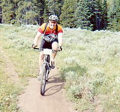 A mountain biker wearing a helmet and colorful jersey rides along a dirt trail surrounded by green foliage and trees. Trestle Bike Park mountain bike trail.