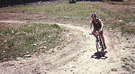 A cyclist riding a mountain bike along a dirt trail in a grassy area. The rider is wearing a helmet and sports attire, navigating a curved path surrounded by sparse vegetation and rocky terrain. Trestle Bike Park mountain bike trail.