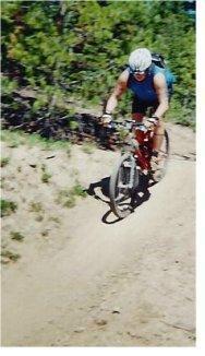 A person riding a mountain bike down a dirt trail surrounded by greenery. The cyclist is wearing a helmet and is focused on navigating the terrain. Trestle Bike Park mountain bike trail.