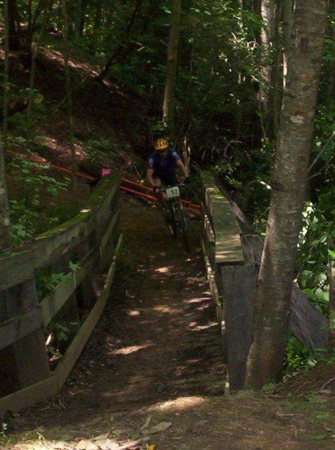 A mountain biker in a yellow helmet navigates a narrow path through a wooded area, crossing a wooden bridge surrounded by trees and greenery. Fontana Village mountain bike trail.